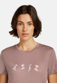 Lilla kortærmet t-shirt med hvide grafiske illustrationer af skiløbere og snowboardere i forskellige poser over brystet.