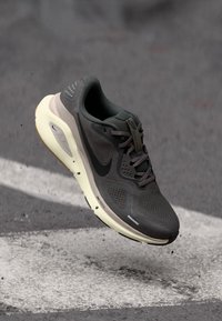 Scarpa da corsa Nike grigio scuro e beige con tomaia in rete e suola air visibile, sospesa sopra una strada asfaltata con segnaletica bianca.