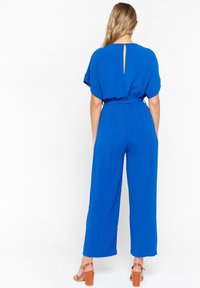 Niebieski kombinezon z krótkim rękawem, elastyczną talią i szerokimi nogawkami. Posiada detal w postaci łezki z tyłu. Gładki, lekki materiał.
