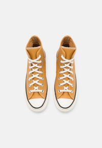 Vysoké tenisky vyrobené z texturované hořčičně žluté kůže, s bílými tkaničkami, bílým gumovým prstem a černými akcenty.