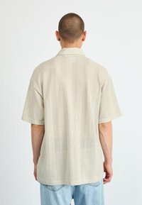 Short-sleeve, light beige polo shirt with a perforated pattern, featuring a fold-down collar and relaxed fit, paired with light blue jeans.