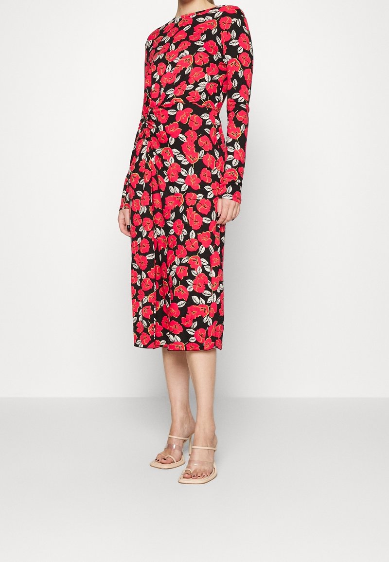 Kvinna iklädd en midiklänning i svart med röd blommig tryck, långa ärmar och beige klacksandaler, stående mot en enfärgad bakgrund.