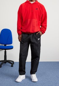Felpa rossa in pile con tasca a marsupio e logo nero, abbinata a pantaloni cargo neri con tasche laterali. Scarpe da ginnastica bianche completano il look.