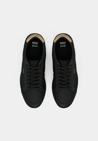 Black sneakers featuring textured fabric, flat laces, and gold accents. Ortholite insole visible on the inside with logo detail.