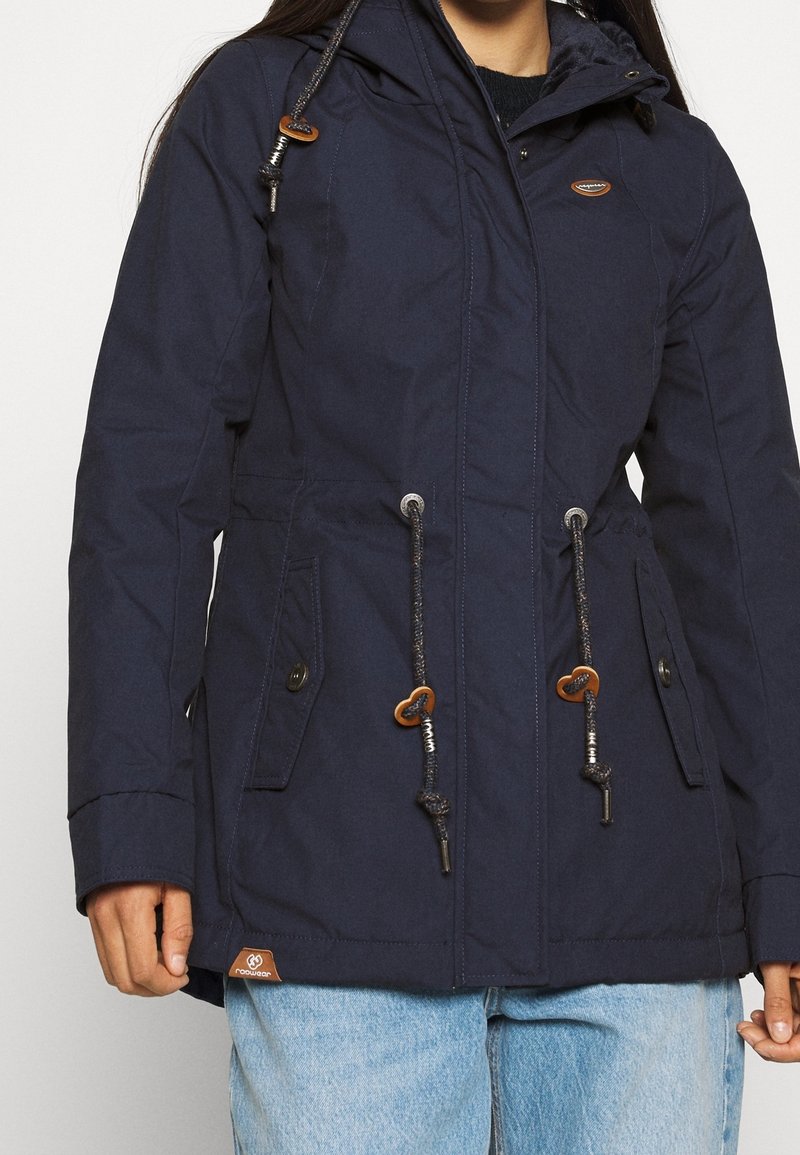 Kobieta w granatowej kurtce z kapturem, z guzikami na kieszeniach, sznurkami z brązowymi zakończeniami oraz jasnoniebieskich dżinsach.