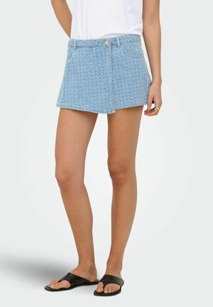 Femme portant un skort en denim bleu clair texturé, un haut blanc et des sandales plates noires à bout ouvert, debout sur fond blanc.