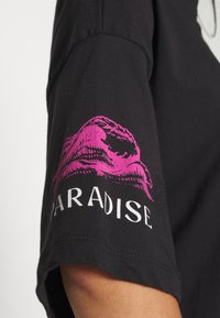 Sort bomulds t-shirt med korte ærmer. Har et pink grafisk motiv af læber og ordet "PARADISE" i hvidt under motivet.