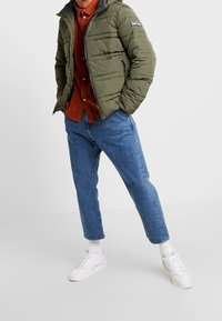 Zielona pikowana kurtka z zamkiem błyskawicznym, bocznymi kieszeniami, szarymi akcentami, noszona na rdzawym podkoszulku, w połączeniu z jasnoniebieskimi dżinsami i białymi sneakersami.