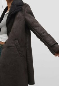 Cappotto in finta pelle marrone con un morbido collo in shearling nero. Presenta una chiusura con un bottone singolo e tasche laterali, evidenziando una vestibilità comoda.