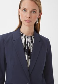 Marineblauwe blazer met een gestructureerd ontwerp, met inkepingen op de revers en een enkele knoop, gelaagd over een gebloemd blouse. Zilveren oorbellen.