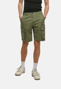 Shorts cargo vert olive avec plusieurs poches, coupe droite, taille élastique et tissu lisse et léger. Portés avec des chaussettes blanches et des baskets.