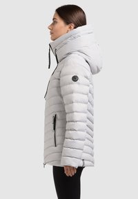 Svetle šedý péřový kabát s vysokým límcem, velkou kapucí, horizontálním prošíváním, bočními zipovými kapsami a černou nášivkou s logem na rukávu.