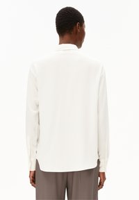 Camicia bianca a maniche lunghe con colletto classico, texture liscia, polsini abbottonati e orlo dritto, indossata sopra dei pantaloni grigi.