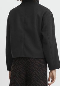 Černá zkrácená bunda s vysokým límcem, strukturovanými rameny a hladkou texturou. Má detaily švů a je kombinována se vzorovanými kalhotami.