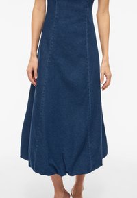 Ženska v temno modri denim midi obleki z oprijetim pasom in balonasto spodnjico, stoji bosonoga na belem ozadju.