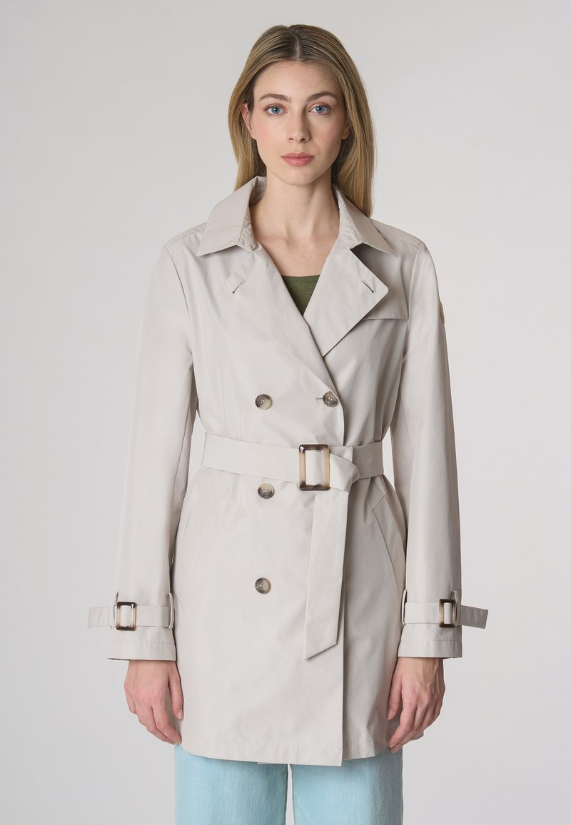 Trench coat beige con design a doppio petto, ampio colletto, chiusure con bottoni, vita con cintura e cinturini regolabili ai polsini. Texture liscia.