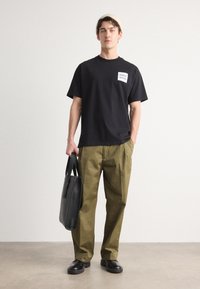 Zwarte katoenen t-shirt met een wit logopatch, olijfgroene broek, zwarte leren schoenen en een zwarte totebag. Gladde textuur en casual pasvorm.
