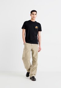 T-shirt noir avec logo jaune, associé à un pantalon cargo beige avec poches latérales ; modèle portant des chaussures noires, se tenant devant un fond blanc.