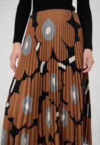 Donna che indossa una gonna plissettata con un grande motivo floreale marrone e blu e un top nero a maniche lunghe, con le mani poggiate sui fianchi.