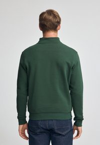 Felpa verde con colletto alto, polsini e bordo a costine. Il tessuto sembra morbido e strutturato. Indossata con jeans blu scuro.