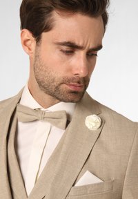 Beige pak met een gestructureerde afwerking, wit shirt en bijpassende vlinderdas. Voorzien van een witte bloemen reversspeld en een zakdoek in de zak van het jasje.