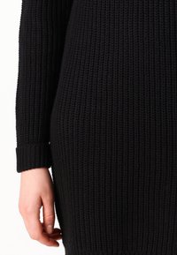 Gros plan d'une main reposant à côté d'une manche et du corps d'un pull en maille noire et épaisse sur un fond clair.