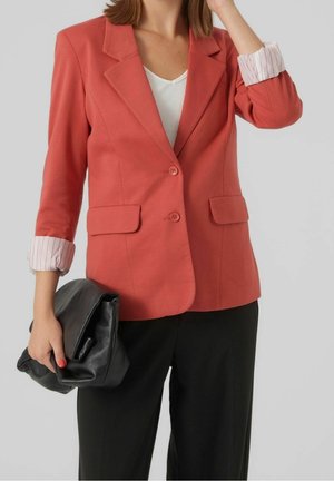 Kvinde iført koralfarvet blazer med op-rullede hvide stribede manchetter, sorte bukser, hvid top, holder en stor sort clutchtaske i venstre hånd.