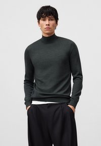 Maglione a collo alto verde scuro realizzato in morbido tessuto lavorato a maglia, caratterizzato da maniche lunghe e una silhouette aderente. Indossato sopra una camicia bianca, abbinato a pantaloni neri.