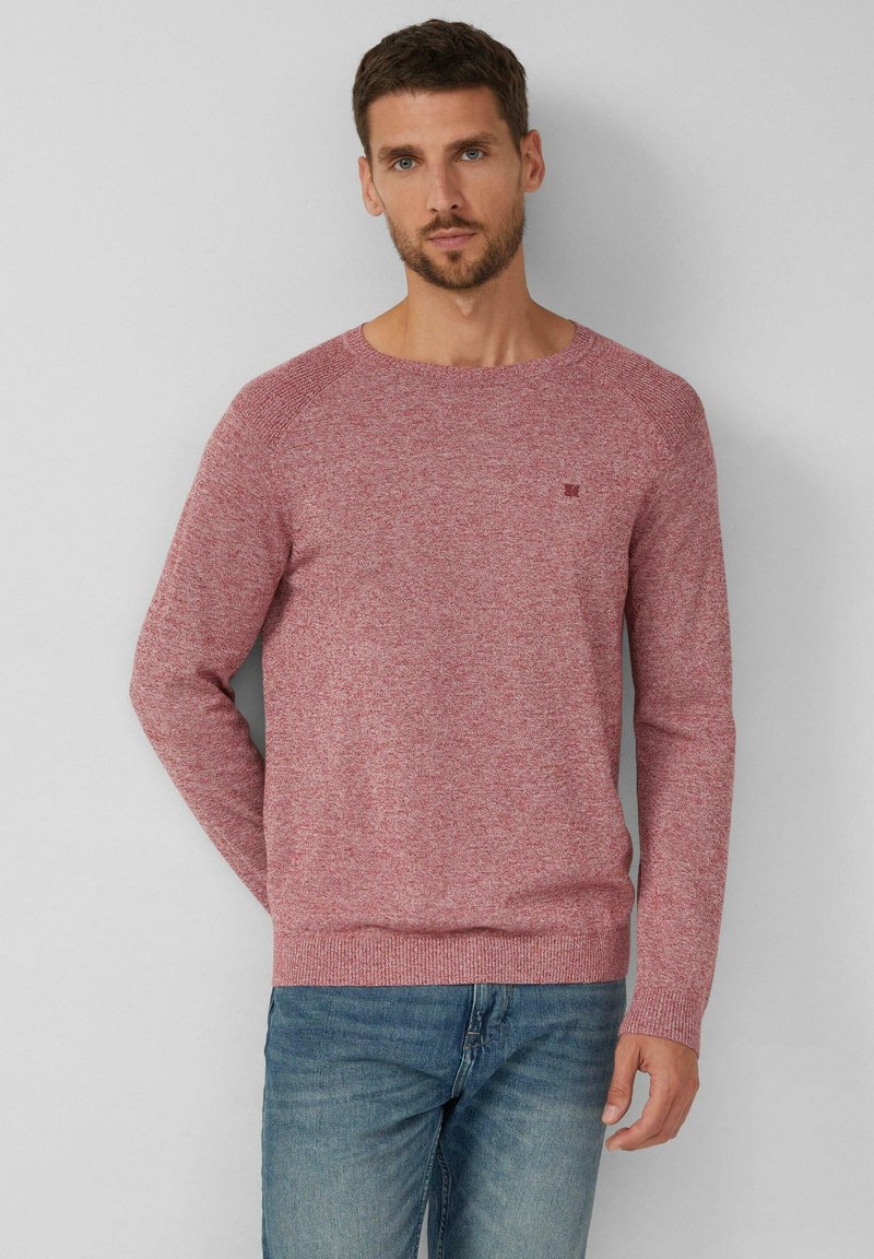 Roter Strickpullover mit langen Ärmeln und runden Halsausschnitt. Abgesetzte Rippbündchen an den Ärmeln und am Saum, kombiniert mit blauen Jeans. Minimales Logodesign.