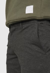 Felpa verde oliva con un'etichetta bianca con la scritta "WZB", abbinata a pantaloni grigio scuro con texture, caratterizzati da tasche frontali e chiusura a bottone.