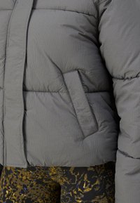 Gros plan d’une veste matelassée grise avec une poche latérale, portée sur un pantalon noir et jaune à motifs.