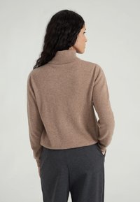 Brauner Rollkragenpullover aus weichem Stoff, mit legerem Schnitt und geripptem Kragen. Kombiniert mit dunkelgrauen, maßgeschneiderten Hosen.