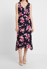 Robe midi florale avec une base marine, ornée de fleurs roses et blanches. Décolleté en V, design sans manches et ourlet fluide.