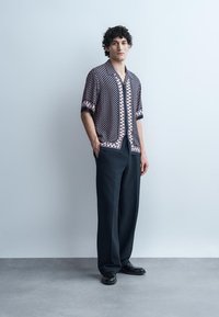 Jonge man staat met handen in zakken, draagt een korte mouw patroonhemd, donkere brede broekspijpen en zwarte schoenen tegen een effen achtergrond.