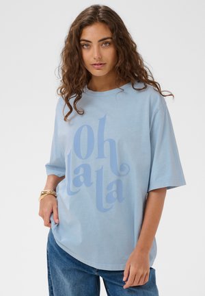 Frau mit lockigem braunem Haar, die ein hellblaues, übergroßes Hemd mit dem Schriftzug "oh la la" und blaue Jeans trägt und vor einem schlichten Hintergrund steht.