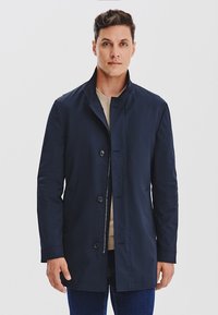 Granatowy płaszcz trench z kołnierzem stójkowym, z przodu z guzikami i gładką fakturą. Nałożony na beżowy sweter, z ciemnymi dżinsami.