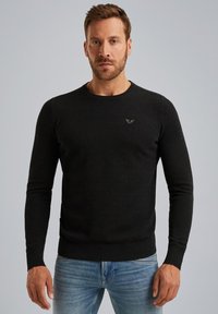 Un pull noir en tricot avec des manches longues, un col rond, un tissu texturé et un petit logo sur la poitrine. Porté avec un jean bleu.
