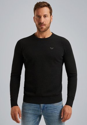 Un pull noir en tricot avec des manches longues, un col rond, un tissu texturé et un petit logo sur la poitrine. Porté avec un jean bleu.
