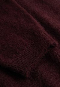 Detailný záber na mäkkú, tmavú bordovú pleteninu s viditeľným rebrovaným okrajom manžety a chlpatou textúrou.