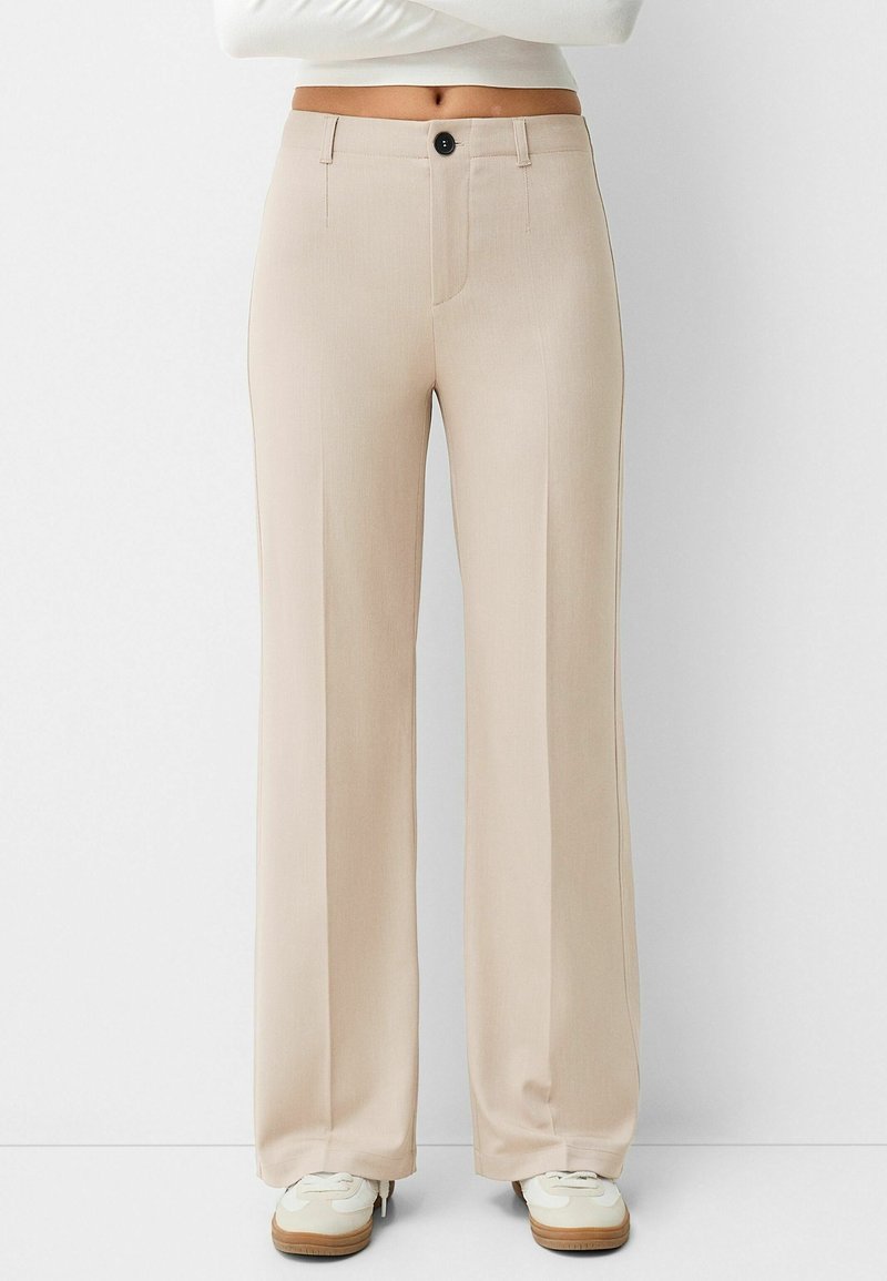 Pantalon beige à jambes larges avec fermeture devant par bouton et zip, porté avec des baskets blanches et un top blanc court visible à la taille.