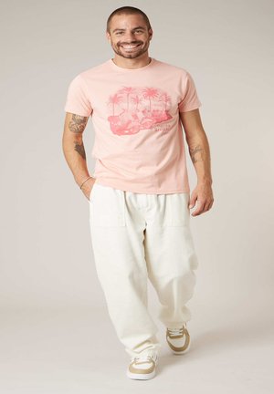 Homme souriant avec des tatouages portant un t-shirt graphique rose Riviera italienne, un pantalon blanc ample et des baskets beige-blanc, marchant en avant.