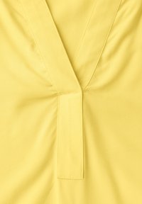 Tissu jaune avec un décolleté en V comportant une large patte rectangulaire et des fronces subtiles autour du col.