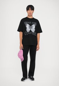 Μαύρη oversized μπλούζα με γραφικό πεταλούδας και κείμενο "Versace Jeans Couture", σε συνδυασμό με μαύρο παντελόνι και ροζ καπέλο. Μαύρα αθλητικά ολοκληρώνουν την εμφάνιση.