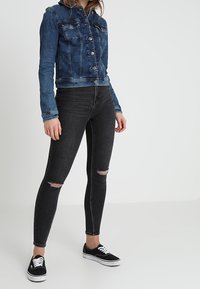 Jeansjacka i mörkblå, två bröstfickor, knappstängning. Svarta skinny jeans med slitningar vid knäna. Svarta sneakers med vita snören.
