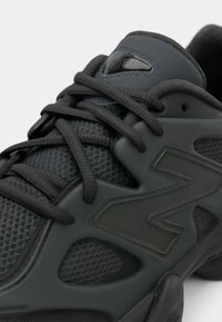 Shoe atlético preto com parte superior em malha respirável, sobreposições texturizadas, logotipo "N" proeminente e atacadores pretos com um colar almofadado.