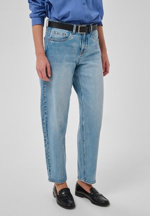 Personne portant un jean bleu clair, une ceinture noire, une chemise à carreaux bleue et des mocassins en cuir noir, debout devant un fond uni.