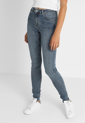 Femme portant un jean bleu ajusté, des baskets blanches et un débardeur blanc court, debout avec une jambe légèrement pliée.