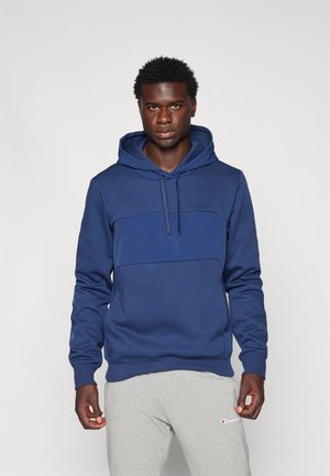 Marineblå hettegenser med flat frontseam og justerbar snorhette. Myk bomullsblanding, kombinert med lysegrå joggebukser.