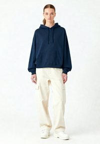 Femeie cu părul brunet prins într-un coadă de cal, purtând un hanorac bleumarin, pantaloni cargo crem și adidași albi, stând în fața unui fundal alb simplu.