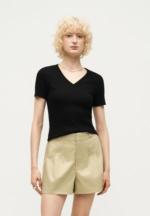 Ξανθός με σγουρά μαλλιά φοράει μαύρο κοντομάνικο μπλουζάκι με V λαιμόκοψη και μπεζ ψηλόμεσα πλισέ σορτς, στέκεται μπροστά σε απλό φόντο.